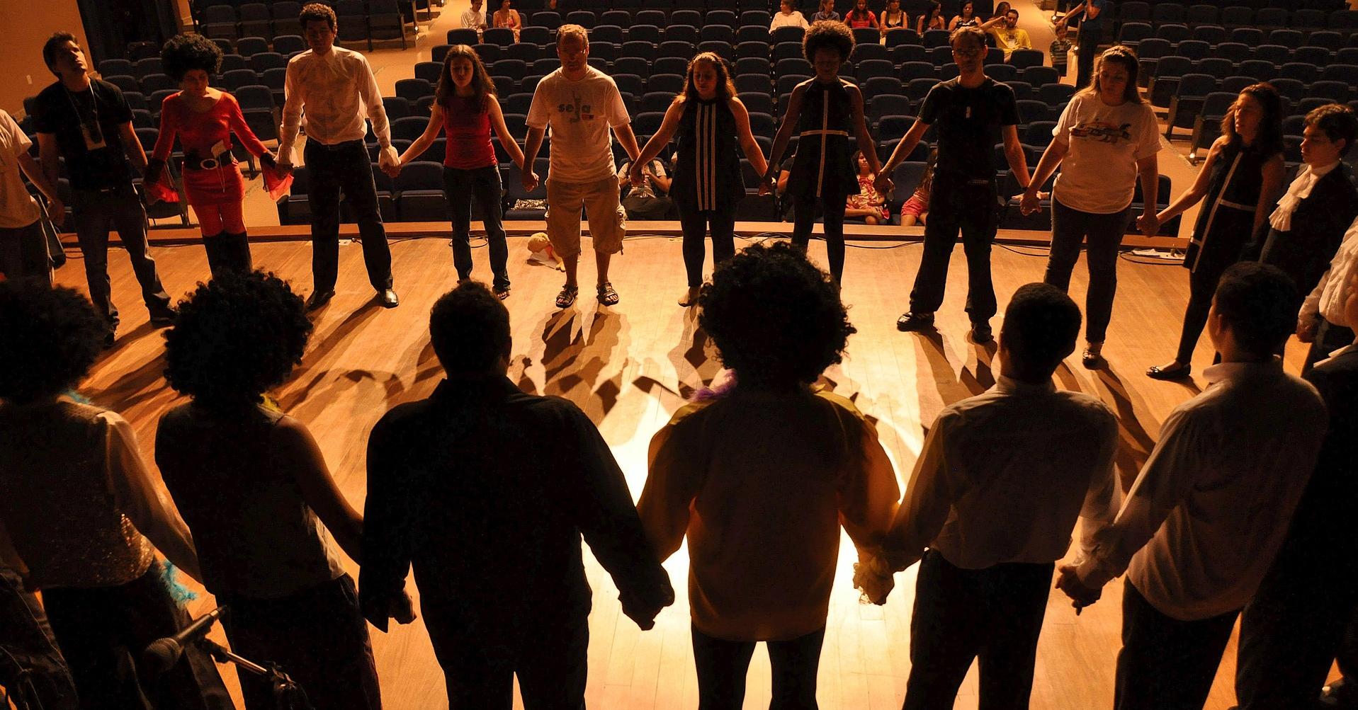 Photo décorative d'un atelier de théâtre où une vingtaine de personnes sont réunies en cercle sur une scène et se tiennent la main