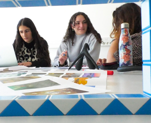 Photo de trois étudiantes assises côte à côté à une table. A gauche de l’image, l’une d’elles est en train de travailler sur ordinateur. Au centre, une autre étudiante discute avec une troisième personne à droite de l’image. Des documents sont posés sur la table devant elles.