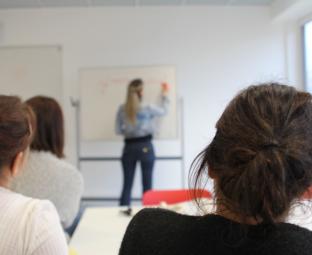 Photo de 3 personnes assises de dos face à un tableau sur lequel une femme est en train d'écrire.