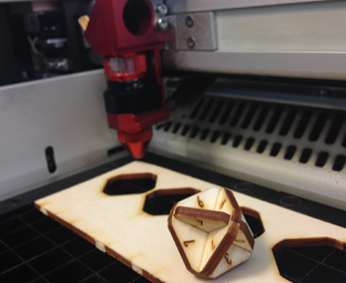 Photo de la découpeuse laser de l'Edulab. Au premier plan, une fine planche de bois dans laquelle viennent d’être découpés quatre petits octogones. Assemblés, ils forment une sorte de dé, posé sur la planche. A l’arrière-plan, le bras du laser, de couleur bordeaux. 