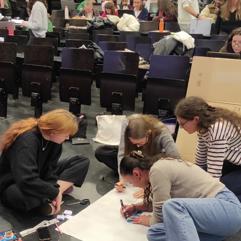 Photo décorative : des étudiantes et étudiants sont rassemblés en groupe dans un amphithéâtre. Certains travaillent assis par terre, tandis que d'autres sont sur des tables.
