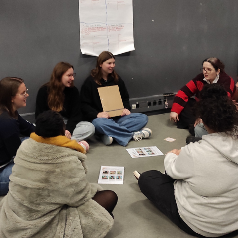 Photo d'étudiantes assises en cercle au sol pour tester un prototype de jeu pédagogique. A leurs pieds, des supports de jeu imprimés et, sur le mur derrière elles, une grande fiche d’évaluation par les pairs, alimentée au fur et à mesure par les groupes qui passent tester le jeu.