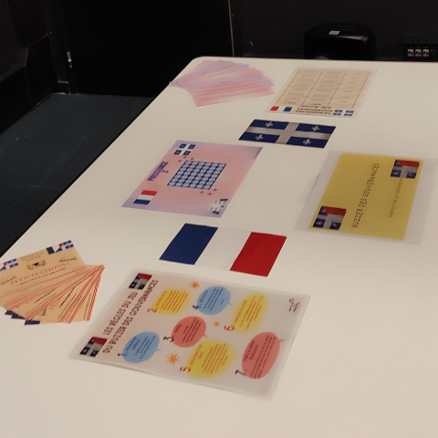 Photo du jeu pédagogique "Le Buzzer des Gouvernances" : divers supports imprimés, notamment des fiches de règles illustrées de bulles colorées, des cartes de félicitations et des plateaux de jeu arborant les drapeaux de la France et du Québec sont disposés sur une table. L'ensemble a été conçu pour une activité pédagogique comparant les systèmes de gouvernance de ces deux régions francophones.