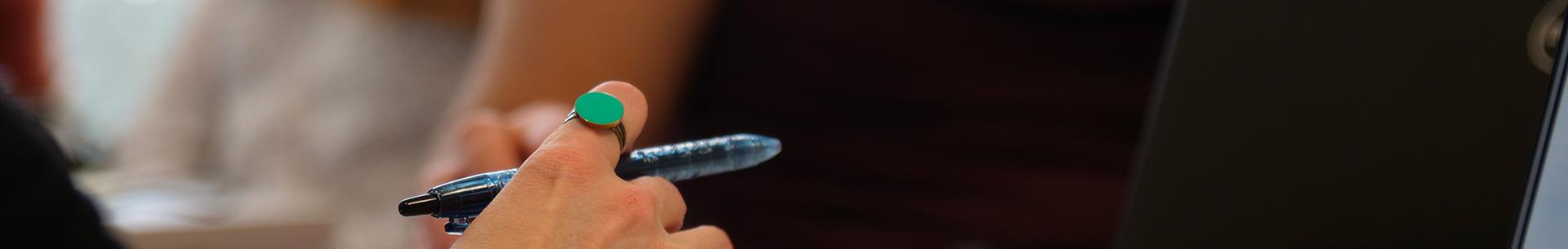 Photo d'une main tenant un stylo, devant un ordinateur ouvert. A l'arrière-plan, des personnes en train de travailler