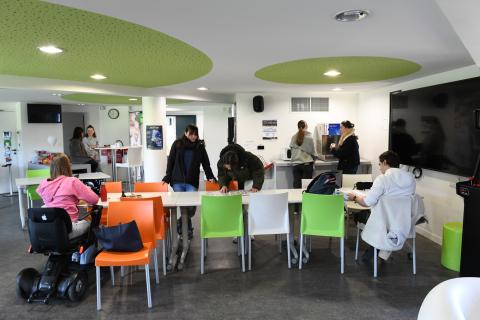 Photo de la salle de convivialité du Ty-Maz' représentant des tables, des chaises, un grand écran vidéo fixé au mur, un distributeur d’eau et un micro-ondes