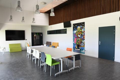 Photo de la salle de pédagogique et culturelle du Ty-Maz' représentant une grande table, des chaises et des fauteuils de différentes couleurs, ainsi qu’un grand écran vidéo fixé au mur